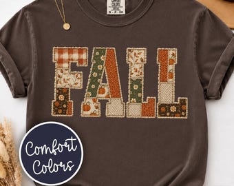 May include: A dark brown short-sleeve t-shirt featuring the word FALL in large block letters. Each letter is filled with a patchwork design of orange, green, and white patterns including pumpkins, leaves, and flowers. The shirt has a crew neckline.