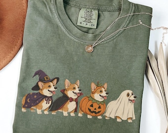 May include: An olive green crew neck t-shirt featuring a graphic of four Corgi dogs dressed in Halloween costumes: a wizard, a vampire, a pumpkin, and a ghost. The shirt has a Comfort Colors label showing size S-P-CH and is made of 100% cotton.