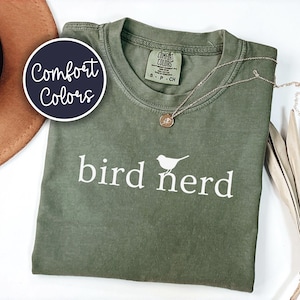 Pu&ograve; includere: Una t-shirt a maniche corte color verde oliva con la scritta bianca bird nerd e una piccola grafica di un uccello sopra la lettera r. La maglia presenta un'etichetta del marchio Comfort Colors sul collo interno e un girocollo.