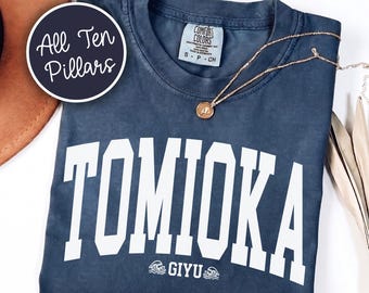 May include: A slate blue short-sleeve t-shirt featuring the white text TOMIOKA in a large arched font and GIYU with small wave icons below. The shirt has a crew neckline and a Comfort Colors brand label at the inner collar. A gold-toned necklace rests on the fabric.