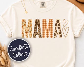 May include: A cream-colored short-sleeve t-shirt featuring the word MAMA in large block letters. Each letter has a different pattern: floral, leopard print, orange plaid, and cow print. Two outlined hearts are positioned to the right of the text.