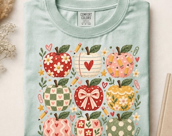 May include: A light green t-shirt with a graphic print of nine stylized apples in various patterns including flowers, hearts, checks, and polka dots, interspersed with small yellow pencils, stars, and paper clips. The neck label reads COMFORT COLORS.