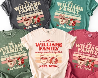 May include: Five short-sleeve crew neck t-shirts in charcoal, teal, olive green, cream, and maroon. Each shirt features a graphic with the text "the WILLIAMS FAMILY making memories together" and "EST. 2026" above a mug and three hanging stockings.