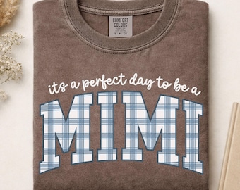 May include: A brown crewneck t-shirt featuring the white text 'its a perfect day to be a' above the word 'MIMI' in large block letters filled with a blue and white plaid pattern. The shirt has a Comfort Colors brand label at the inner neckline.