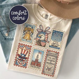 May include: Cream-colored t-shirt featuring a collection of vintage-style postage stamps with patriotic designs. The stamps depict the Liberty Bell, an eagle, fireworks, a ship, a top hat, and Independence Hall. The shirt also has the text "Comfort Colors".