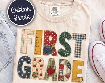 May include: A cream-colored t-shirt featuring the text FIRST GRADE in large, patchwork-style letters. The letters are decorated with various patterns including plaid, polka dots, stars, and school-themed graphics like apples, pencils, and a ruler.