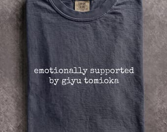May include: A dark gray short-sleeved t-shirt with a crew neckline. The shirt features white text printed on the chest that reads, "emotionally supported by giyu tomioka." A tan brand label is visible inside the collar of the garment.