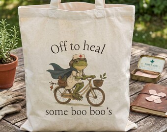 May include: A cream-colored canvas tote bag featuring a graphic of a frog dressed as a nurse riding a bicycle. The frog wears a nurse cap and cape. The text above and below the illustration reads, Off to heal some boo boo's.