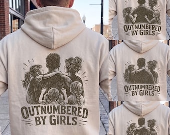 May include: A beige hooded sweatshirt featuring a dark brown graphic on the back. The illustration depicts a central figure surrounded by three children and one adult, with the text "OUTNUMBERED BY GIRLS" printed in a bold, stylized font below.