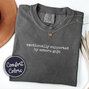 May include: Dark gray Comfort Colors t-shirt with the text "emotionally supported by satoru gojo" in white. A silver necklace with a small pendant is draped across the shirt. A Comfort Colors logo is visible on the bottom left.
