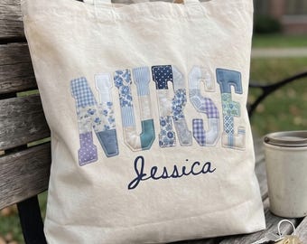 May include: A cream-colored canvas tote bag featuring the word NURSE in large, block-style letters made from various blue and white patterned fabric patches. Below the word, the name Jessica is embroidered in a dark blue cursive script font.