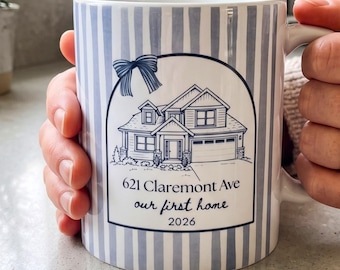 May include: A white ceramic mug featuring a light blue vertical striped pattern. The front displays a dark blue line drawing of a house with a bow above it, and the text 621 Claremont Ave, our first home, and the year 2026 printed below.