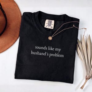 May include: A black short-sleeved t-shirt with a crew neckline featuring the white text "sounds like my husband's problem" printed on the chest. The shirt has a Comfort Colors brand label inside the collar and is displayed with a gold necklace.