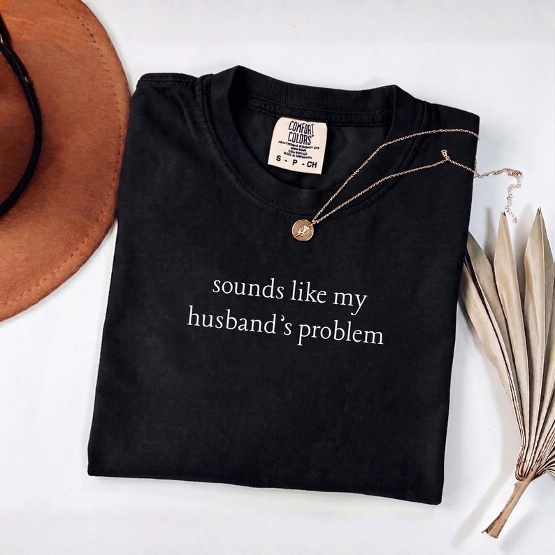 May include: A black short-sleeved t-shirt with a crew neckline featuring the white text "sounds like my husband's problem" printed on the chest. The shirt has a Comfort Colors brand label inside the collar and is displayed with a gold necklace.