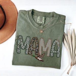 Peut inclure: Un t-shirt &agrave; manches courtes vert olive avec le mot MAMA imprim&eacute; en grandes lettres. Les lettres pr&eacute;sentent des motifs vari&eacute;s, notamment des imprim&eacute;s l&eacute;opard, vache et des rayures verticales. Un canard colvert est illustr&eacute; sous le texte.