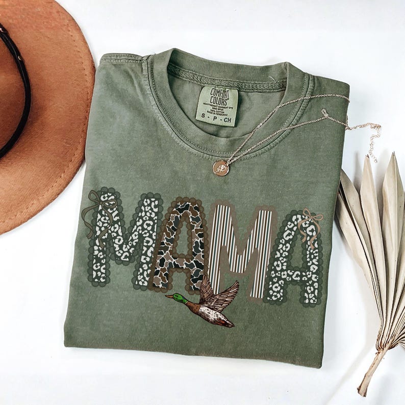 Peut inclure: Un t-shirt &agrave; manches courtes vert olive avec le mot MAMA imprim&eacute; en grandes lettres. Les lettres pr&eacute;sentent des motifs vari&eacute;s, notamment des imprim&eacute;s l&eacute;opard, vache et des rayures verticales. Un canard colvert est illustr&eacute; sous le texte.