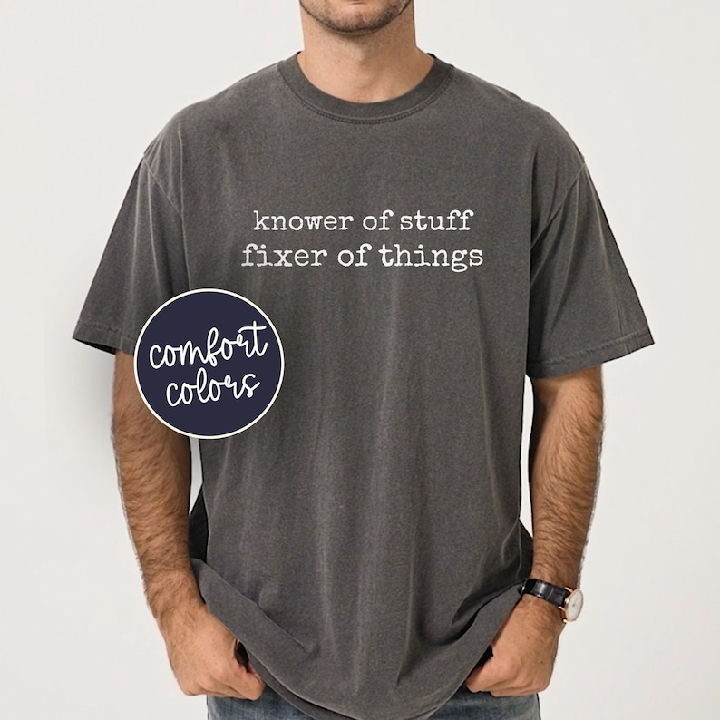 Op de afbeelding: Donkergrijs t-shirt met de witte tekst "knower of stuff fixer of things". Een marineblauwe cirkel met de woorden "comfort colors" staat aan de linkerkant van het shirt. Het shirt heeft korte mouwen en een ronde hals.