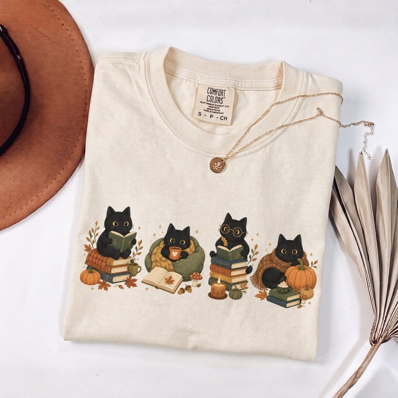 May include: A cream-colored t-shirt with a graphic print of four black cats engaged in autumn activities. The cats are shown reading books, drinking from a mug, and sitting near pumpkins and fall foliage. The shirt features a Comfort Colors brand label.