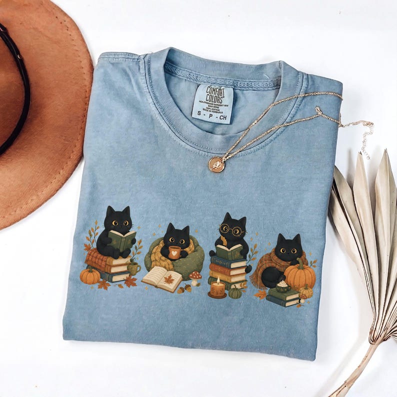 May include: A light blue t-shirt featuring a graphic of four black cats engaged in autumn activities. The cats are depicted reading books, drinking from mugs, and sitting near pumpkins, fall leaves, and candles. The shirt has a Comfort Colors label.