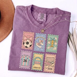 May include: A purple Comfort Colors t-shirt featuring six postage stamp designs with soccer-themed illustrations. The stamps include a soccer ball, cleats, a trophy, a jersey, a goal, and the text "World Cup 2026". A silver necklace with a pendant is also visible.