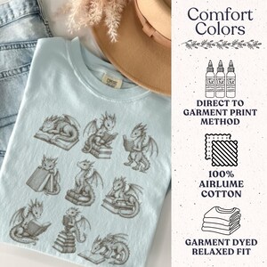 K&ouml;nnte beinhalten: Ein hellblaues T-Shirt mit einem Grafikdruck von neun kleinen Drachen, die mit B&uuml;chern interagieren. Der Text Comfort Colors ist auf dem inneren Nackenetikett sichtbar. Auf der rechten Seite steht: Comfort Colors, Direct to Garment Print Method, 100% Airlume Cotton und Garment Dyed Relaxed Fit.