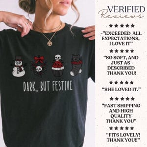 Op de afbeelding: Een zwart t-shirt met een afbeelding van een sneeuwpop, maretak met een schedel, een cupcake met een schedel en een kat in een kerstsok. De tekst luidt "DARK, BUT FESTIVE". De afbeelding bevat de kleuren zwart, wit en rood.