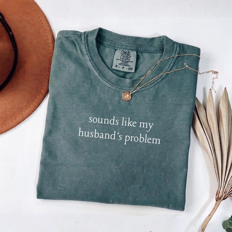 May include: A slate green short-sleeve t-shirt with a crew neckline and a Comfort Colors brand label at the inner collar. The front features the white text "sounds like my husband's problem" printed in a simple, lowercase sans-serif font.