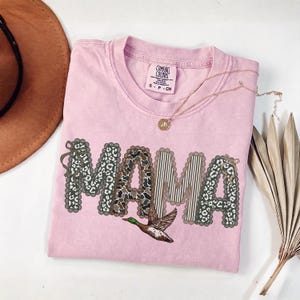 Peut inclure: Un t-shirt rose clair &agrave; manches courtes et col rond. L'avant pr&eacute;sente le mot MAMA en grandes lettres aux bords festonn&eacute;s, remplies de motifs l&eacute;opard et ray&eacute;s. Un graphique de canard colvert est positionn&eacute; sous le texte.
