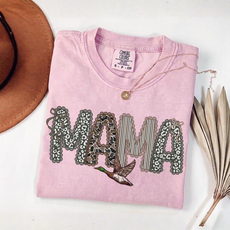Peut inclure: Un t-shirt rose clair &agrave; manches courtes et col rond. L'avant pr&eacute;sente le mot MAMA en grandes lettres aux bords festonn&eacute;s, remplies de motifs l&eacute;opard et ray&eacute;s. Un graphique de canard colvert est positionn&eacute; sous le texte.