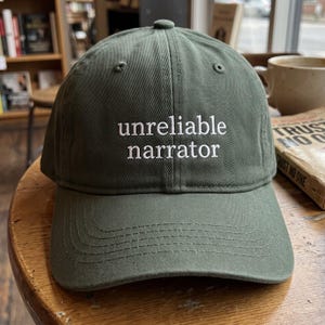 May include: An olive green baseball cap featuring the text "unreliable narrator" embroidered in white thread on the front panel. The cap has a structured crown, a curved brim with tonal stitching, and a button at the top center of the crown.