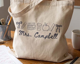 May include: A beige canvas tote bag featuring a dark blue graphic print of a bow, pencil, stack of books, coffee cup with a heart, apple, ruler, and another bow. Below the icons, the text Mrs. Campbell is printed in a dark blue script font.