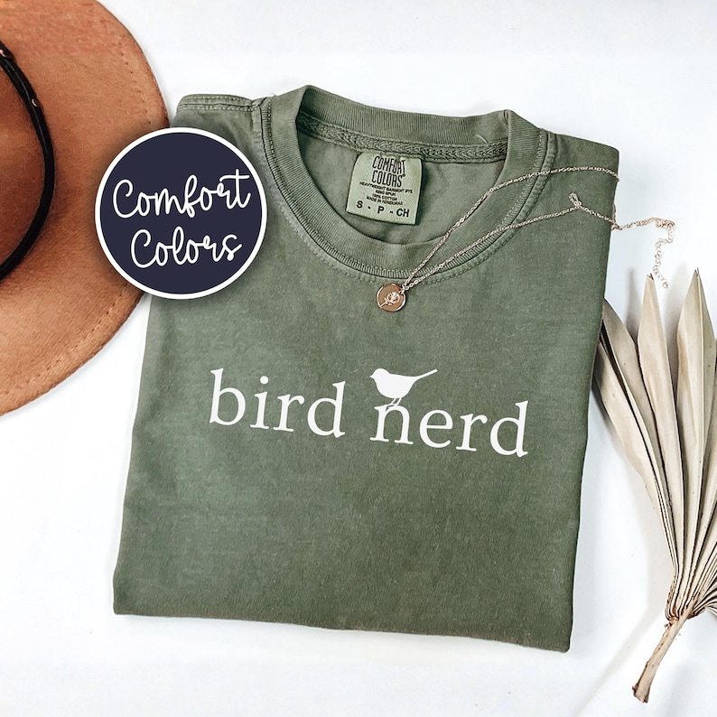 Pu&ograve; includere: Una t-shirt a maniche corte color verde oliva con la scritta bianca bird nerd e una piccola grafica di un uccello sopra la lettera r. La maglia presenta un'etichetta del marchio Comfort Colors sul collo interno e un girocollo.
