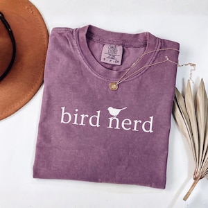 Pu&ograve; includere: Una t-shirt a maniche corte color malva con scollo rotondo, caratterizzata dalla scritta bianca "bird nerd" e da una piccola grafica di un uccello centrata sul petto. La maglietta presenta un'etichetta Comfort Colors nel colletto con taglia S.