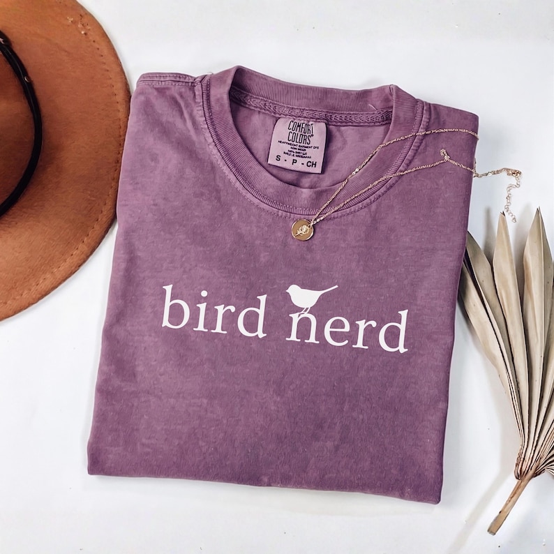 Pu&ograve; includere: Una t-shirt a maniche corte color malva con scollo rotondo, caratterizzata dalla scritta bianca "bird nerd" e da una piccola grafica di un uccello centrata sul petto. La maglietta presenta un'etichetta Comfort Colors nel colletto con taglia S.