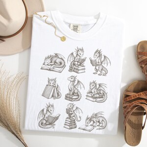 K&ouml;nnte beinhalten: Ein wei&szlig;es T-Shirt mit einem schwarzen Grafikdruck im Tintenstil, der neun kleine Drachen in verschiedenen Posen mit B&uuml;chern zeigt, darunter schlafend auf, sitzend auf oder lesend in B&uuml;chern. Das Shirt hat ein Comfort Colors Etikett am Hals.