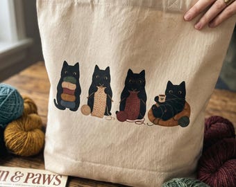 May include: A natural-colored canvas tote bag featuring a graphic print of four black cats engaged in knitting activities. The cats are shown holding yarn, knitting with needles, wearing glasses, and relaxing with a mug, all rendered in a flat illustration style.