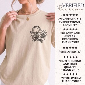 May include: A beige short-sleeved t-shirt featuring a black line art graphic of a long-haired anime character on the left chest. The image includes text reviews with five-star ratings stating: EXCEEDED ALL EXPECTATIONS, SO SOFT, SHE LOVED IT.