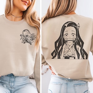 May include: A beige crewneck sweatshirt featuring a small black line art graphic of an anime character on the front left chest and a large black line art graphic of the same character on the back, depicting a girl with long hair and a bamboo muzzle.