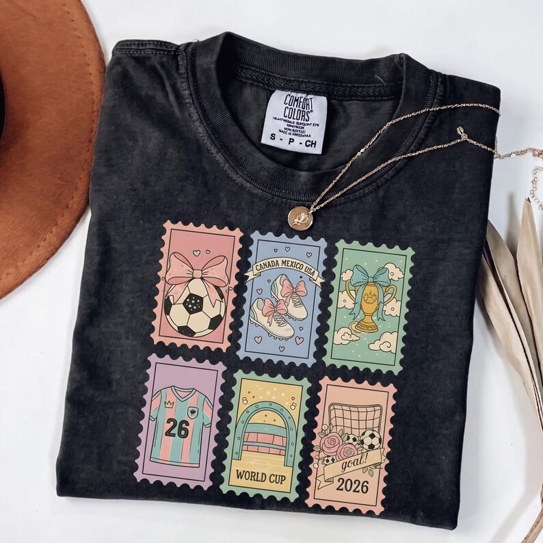 May include: Dark gray Comfort Colors t-shirt with a graphic of soccer-themed postage stamps. The stamps feature a soccer ball, shoes, a trophy, a jersey, a goal, and the text "CANADA MEXICO USA", "WORLD CUP", and "goal! 2026". A gold necklace is draped across the shirt.