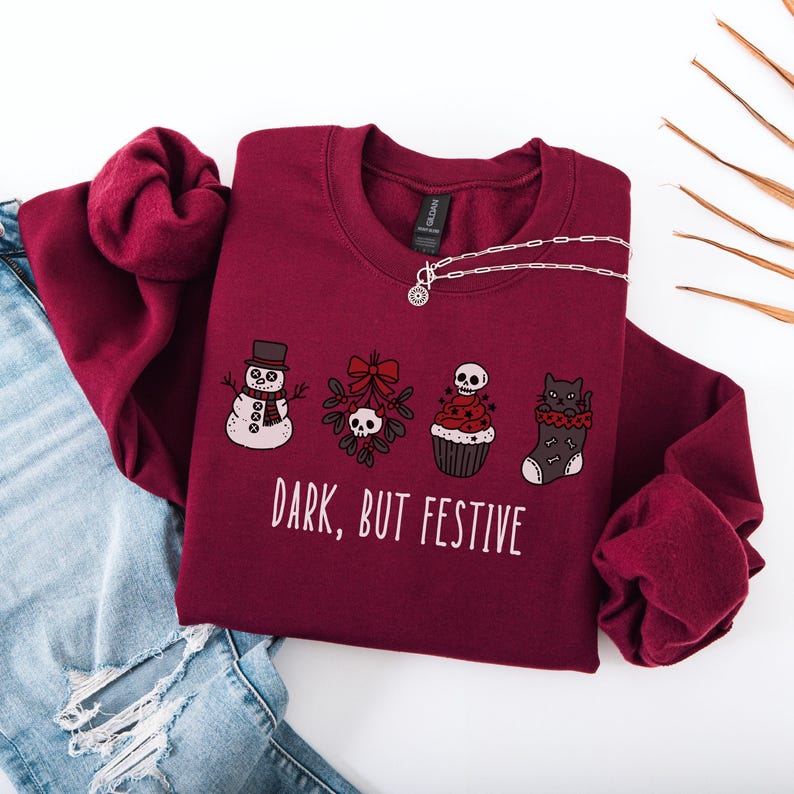 Op de afbeelding: Een bordeauxrode sweater met ronde hals en een afbeelding van een sneeuwpop, maretak met een schedel, een cupcake met een schedel en een zwarte kat in een kerstsok, met de tekst "DARK, BUT FESTIVE" in het wit eronder gedrukt.