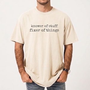 Op de afbeelding: Een beige t-shirt met de tekst "knower of stuff fixer of things" in het zwart gedrukt. Het shirt heeft korte mouwen en een ronde hals. Het shirt is gemaakt van een zacht, comfortabel materiaal.