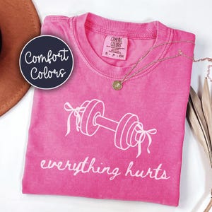 May include: A pink t-shirt with the words "everything hurts" and a dumbbell graphic. The shirt has a crew neck and short sleeves. A gold necklace with a pendant is on the shirt. The shirt is from Comfort Colors.