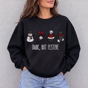 Op de afbeelding: Een zwart sweatshirt met lange mouwen en een grafische print van een sneeuwpop, maretak met een schedel, een cupcake met een schedel als topping en een kat in een kerstsok. De tekst "DARK, BUT FESTIVE" staat in het wit onder de afbeeldingen.