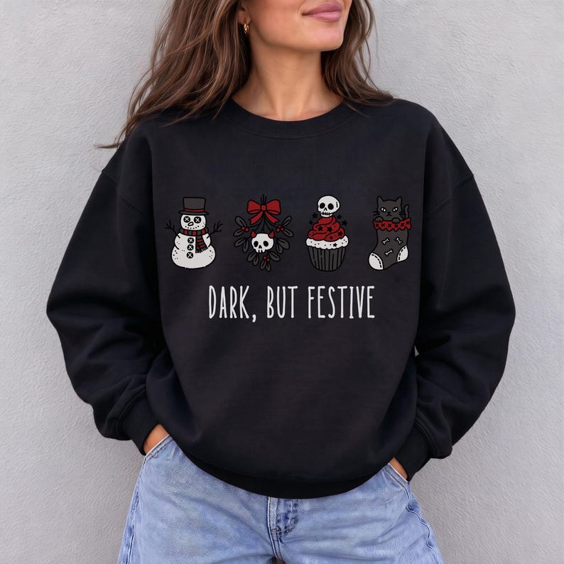 Op de afbeelding: Een zwart sweatshirt met lange mouwen en een grafische print van een sneeuwpop, maretak met een schedel, een cupcake met een schedel als topping en een kat in een kerstsok. De tekst "DARK, BUT FESTIVE" staat in het wit onder de afbeeldingen.