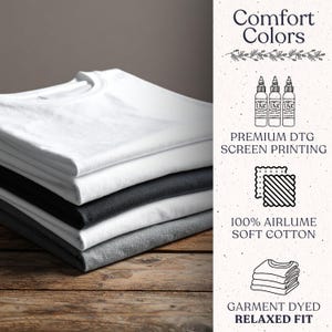 Op de afbeelding: Een stapel opgevouwen t-shirts in wit, zwart en grijs op een houten oppervlak. De afbeelding bevat de tekst "Comfort Colors", "Premium DTG Screen Printing", "100% Airlume Soft Cotton" en "Garment Dyed Relaxed Fit".