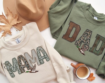 Op de afbeelding: Een crèmekleurig sweatshirt met het woord MAMA in patronenletters en een eendafbeelding, en een olijfgroen sweatshirt met het woord DAD in patronenletters en een eendafbeelding, vergezeld door een beige hoed en een mok koffie.