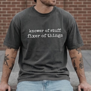 Op de afbeelding: Donkergrijs T-shirt met de witte tekst "knower of stuff fixer of things". Het shirt heeft een ronde hals en korte mouwen. De persoon draagt een blauwe spijkerbroek.