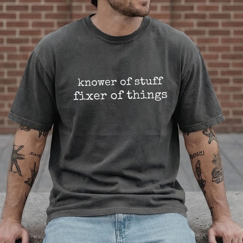 Op de afbeelding: Donkergrijs T-shirt met de witte tekst "knower of stuff fixer of things". Het shirt heeft een ronde hals en korte mouwen. De persoon draagt een blauwe spijkerbroek.