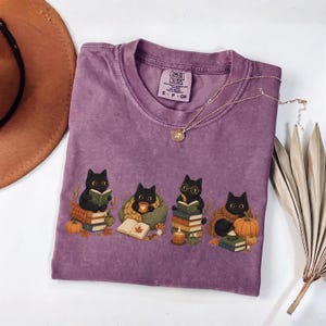 May include: A purple short-sleeve t-shirt featuring a graphic of four black cats engaged in autumn activities, including reading books, drinking from a mug, and sitting near pumpkins and fall foliage. The shirt has a Comfort Colors brand label.