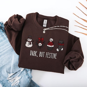 Op de afbeelding: Een donkerbruine sweater met ronde hals en een grafische print van een sneeuwpop, maretak met een schedel, een cupcake met een schedel als topping en een kat in een kerstsok. De tekst "DARK, BUT FESTIVE" staat in witte letters.